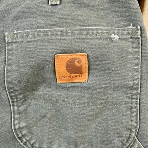 Pu&ograve; includere: Primo piano di un paio di pantaloni da lavoro verdi con una toppa in pelle marrone su cui &egrave; scritto "Carhartt".