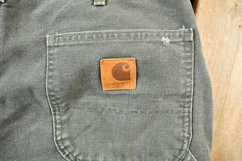 Pu&ograve; includere: Primo piano di un paio di pantaloni da lavoro verdi con una toppa in pelle marrone su cui &egrave; scritto "Carhartt".