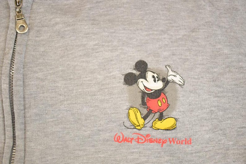 Op de afbeelding: Grijze sweatshirt met ritssluiting met een Mickey Mouse-afbeelding. Het stripfiguur is afgebeeld in zijn klassieke outfit met rode shorts en gele schoenen. De woorden "Walt Disney World" zijn in het rood gedrukt.