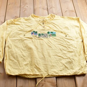 Könnte beinhalten: Ein gelbes Langarmshirt mit einem Kordelzug am Ausschnitt und einem kleinen gestickten Design auf der Vorderseite. Das Design zeigt eine Reihe kleiner Szenen mit Menschen und Bäumen.
