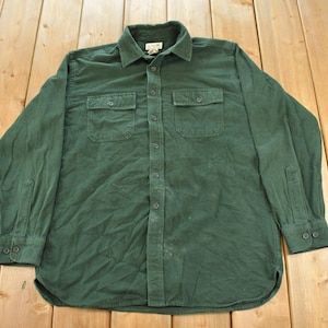 May include: A long-sleeve button-down shirt in a solid dark green color. The shirt features a standard collar, a front button placket, and two chest pockets with buttoned flaps. The cuffs are also buttoned, and the hem has a rounded shape.