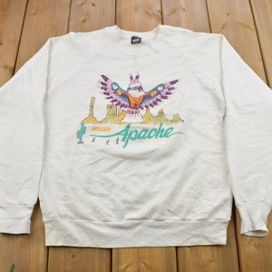 Peut inclure: Sweat-shirt blanc à col rond avec un motif Arizona Apache coloré. Le motif représente un oiseau stylisé aux ailes déployées au-dessus d'un paysage désertique, avec les mots "Arizona Apache" en turquoise.