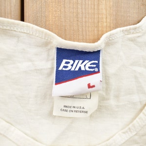 May include: White tank top with a blue tag that says "BIKE" in white letters. The tag also says "Made in U.S.A. Care on reverse."