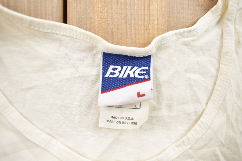 May include: White tank top with a blue tag that says "BIKE" in white letters. The tag also says "Made in U.S.A. Care on reverse."