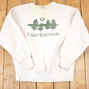 May include: A light gray sweatshirt with the words "CAMP KNUTSON" printed in green. Above the text is a green graphic of trees and a circle. The sweatshirt has long sleeves and a crew neck.