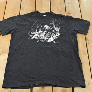 May include: Black t-shirt featuring a white graphic of sailboats on the water. The design includes several sailboats with their sails filled, and a sun or moon in the background. The shirt is made of a soft material.