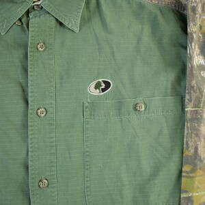 May include: Green button-down shirt with two chest pockets and a collared neckline. The shirt features a camouflage pattern on the sleeves and a small embroidered logo on the chest. The shirt is made of a durable fabric.