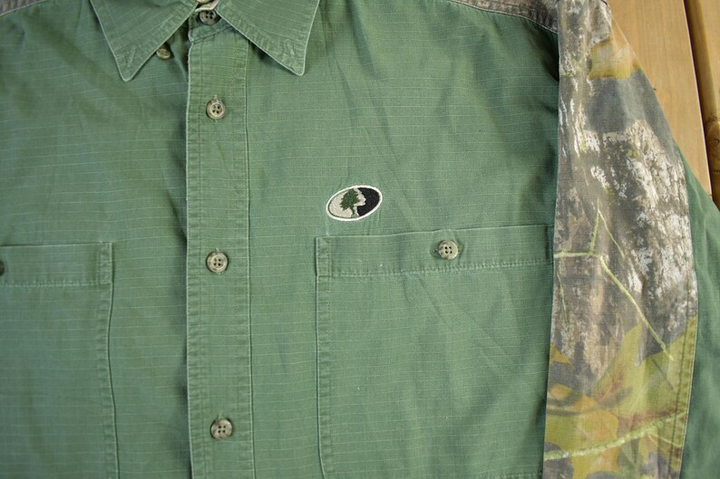 May include: Green button-down shirt with two chest pockets and a collared neckline. The shirt features a camouflage pattern on the sleeves and a small embroidered logo on the chest. The shirt is made of a durable fabric.