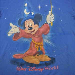 Pu&ograve; includere: Una maglietta blu con una grafica di Topolino vestito da mago con una tunica rossa e un cappello a punta. La grafica include anche stelle e il testo "Walt Disney World".