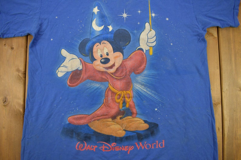 Pu&ograve; includere: Una maglietta blu con una grafica di Topolino vestito da mago con una tunica rossa e un cappello a punta. La grafica include anche stelle e il testo "Walt Disney World".