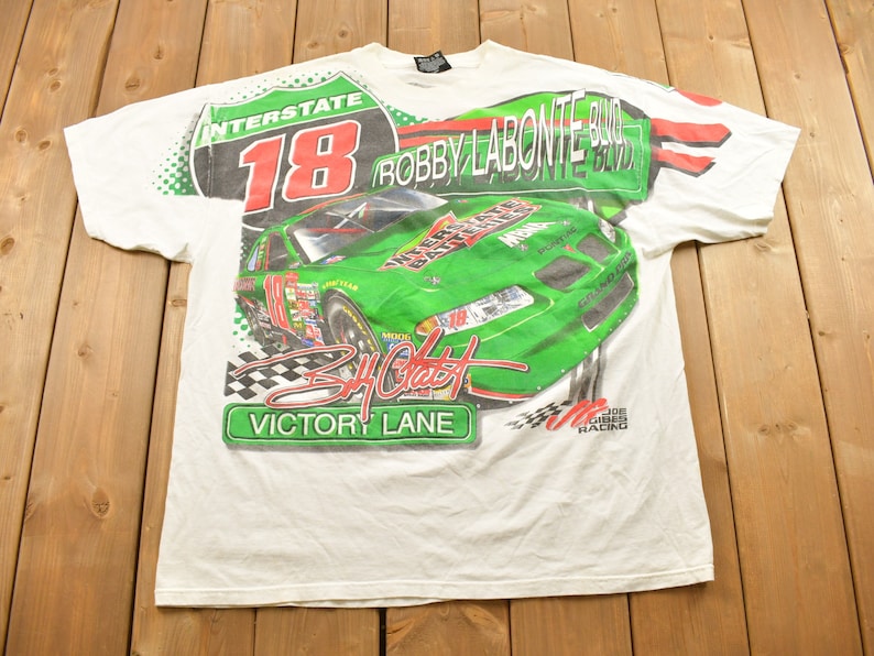 May include: White t-shirt featuring a green and red graphic print of a racing car, the number 18, and the text "Interstate 18 Bobby Labonte Blvd. Victory Lane Joe Gibbs Racing".