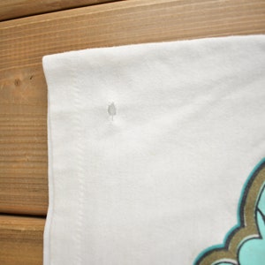 May include: White t-shirt with a small hole near the shoulder seam. The shirt has a graphic design on the front, featuring a turquoise and gold design.