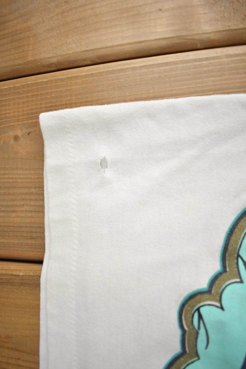 May include: White t-shirt with a small hole near the shoulder seam. The shirt has a graphic design on the front, featuring a turquoise and gold design.