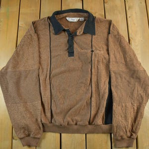 May include: Brown long-sleeved shirt with a black collar and black accents. The shirt has a textured pattern, a three-button placket, and a small pocket. The sleeves have a patch design. The shirt is made of a soft material.