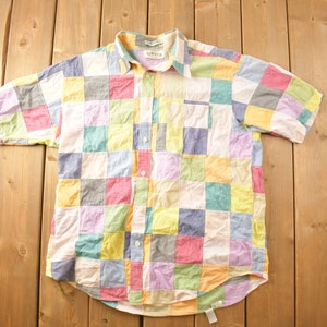 Peut inclure: Une chemise à manches courtes à boutons en patchwork avec une variété de couleurs, notamment le rose, le jaune, le bleu et le vert. La chemise a une poche plaquée sur le côté gauche et une étiquette portant l'inscription "Orvis" sur le col.