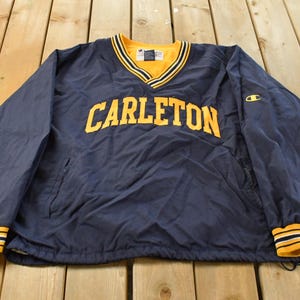 May include: Navy blue pullover jacket with the word "CARLETON" in yellow block letters across the chest. The jacket has a yellow and black striped collar and cuffs. A small Champion logo is on the right sleeve.