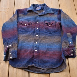 May include: A long-sleeved, button-up shirt with a faded ombre pattern in shades of blue, brown, and purple. The shirt has two chest pockets with button closures and a single button on each cuff.