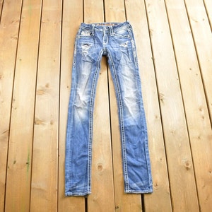 K&ouml;nnte beinhalten: Eine blaue Jeanshose mit Destroyed-Details und wei&szlig;en N&auml;hten. Die Jeans liegt flach auf einer Holzoberfl&auml;che.