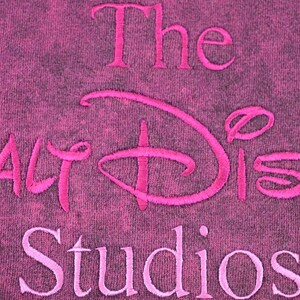 May include: A dark magenta fabric with a mottled, distressed texture featuring the embroidered text The Walt Disney Studios in a bright pink thread. The embroidery is centered, with the word The above, the logo in the middle, and Studios below.