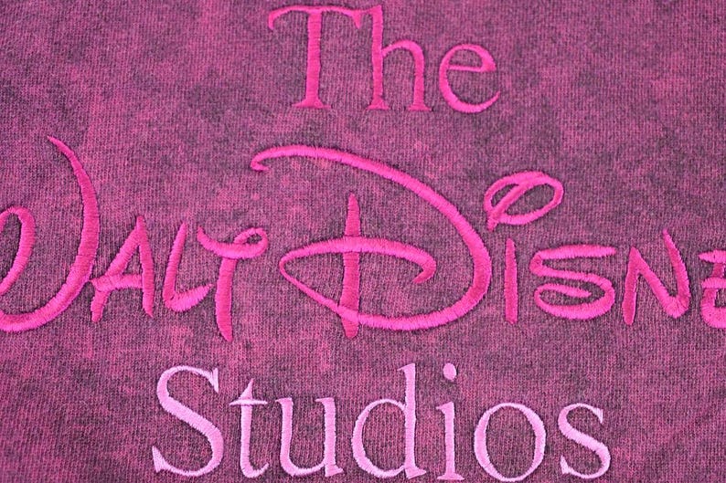 May include: A dark magenta fabric with a mottled, distressed texture featuring the embroidered text The Walt Disney Studios in a bright pink thread. The embroidery is centered, with the word The above, the logo in the middle, and Studios below.