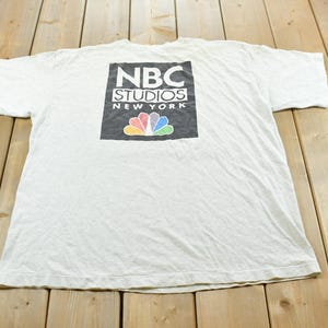 May include: A white short-sleeve t-shirt featuring a black rectangular graphic on the back. The graphic displays the text NBC STUDIOS NEW YORK in white lettering above the iconic multicolored peacock logo with red, orange, yellow, green, blue, and violet.