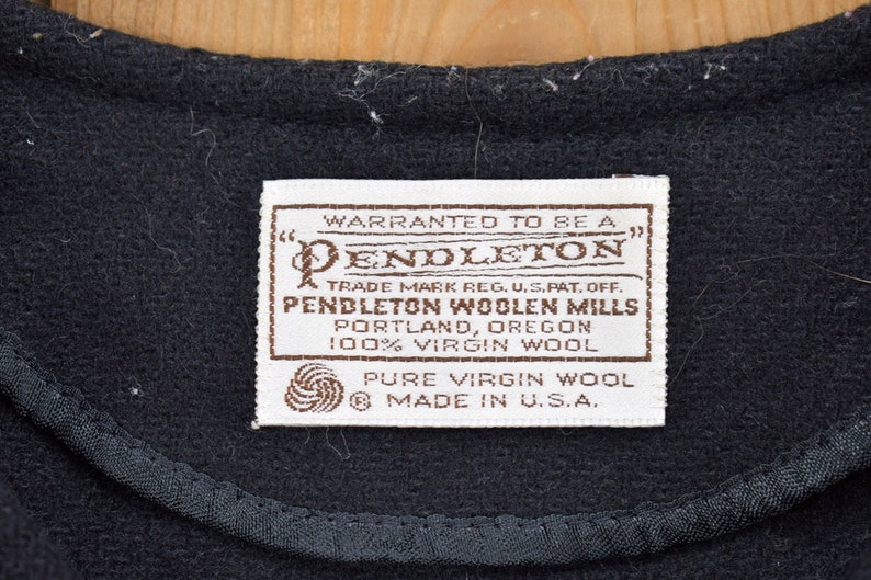 K&ouml;nnte beinhalten: Nahaufnahme eines schwarzen Stoffes mit einer gen&auml;hten Kante und einem rechteckigen Etikett. Das Etikett lautet "PENDLETON WOOLEN MILLS, PORTLAND, OREGON, 100% VIRGIN WOOL, PURE VIRGIN WOOL, MADE IN U.S.A."