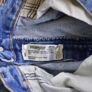 May include: Close-up of a blue denim Wrangler jeans waistband with a white tag that reads "Wrangler 13MWZGK 28 x 36 Machine wash hot and inside out. Tumble twice, times with like colors. Tumble dry. Made in Mexico of US fabric. 100% cotton."