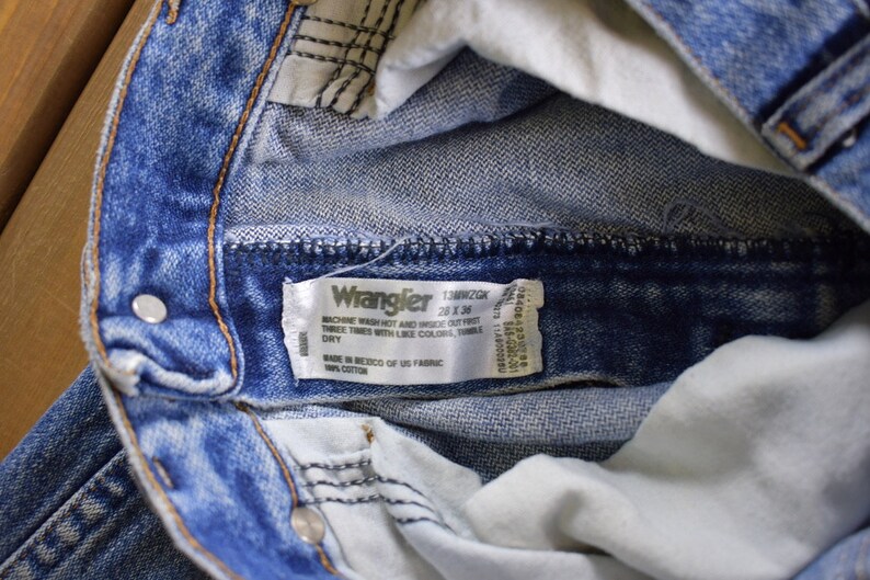 May include: Close-up of a blue denim Wrangler jeans waistband with a white tag that reads "Wrangler 13MWZGK 28 x 36 Machine wash hot and inside out. Tumble twice, times with like colors. Tumble dry. Made in Mexico of US fabric. 100% cotton."