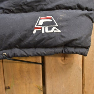 May include: Black puffer jacket with a Fila logo embroidered on the front. The logo is a white triangle with a red stripe and a white "F" inside. The text "FILA" is in white below the triangle.