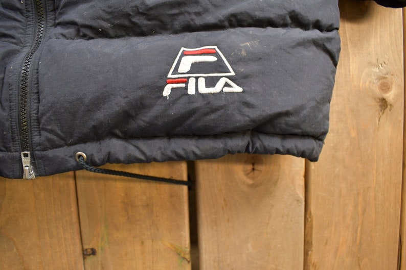 May include: Black puffer jacket with a Fila logo embroidered on the front. The logo is a white triangle with a red stripe and a white "F" inside. The text "FILA" is in white below the triangle.