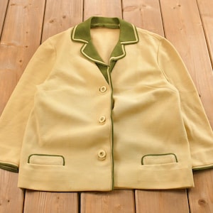 Puede incluir: Un blazer amarillo claro vintage con cuello y ribete verdes. El blazer tiene tres botones y dos bolsillos delanteros con ribete verde. La chaqueta está sobre una superficie de madera.