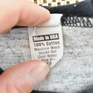 May include: Close-up of a tag from a garment, held by a hand. The tag reads "Made in USA" and provides washing instructions: 100% Cotton, Machine Wash Inside Out, Cool Water, Drip Dry.