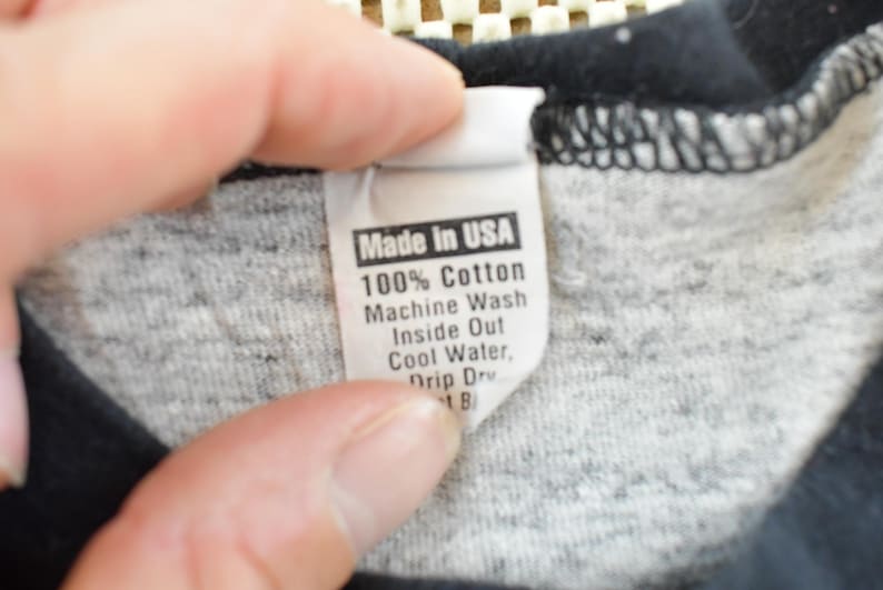 May include: Close-up of a tag from a garment, held by a hand. The tag reads "Made in USA" and provides washing instructions: 100% Cotton, Machine Wash Inside Out, Cool Water, Drip Dry.