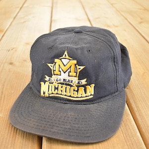 Peut inclure: Casquette de baseball noire avec une visière incurvée. L'avant de la casquette présente un motif brodé or et blanc avec le mot "MICHIGAN" et la phrase "GO BLUE". La casquette est en tissu résistant.