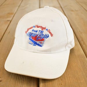 Könnte beinhalten: Weiße Baseballkappe mit gesticktem Text und einer Flugzeuggrafik. Der Text lautet "Sullivan Hartogh Davis Post 730 Honor Flight" in Rot und Blau. Die Kappe liegt auf einer Holzoberfläche.