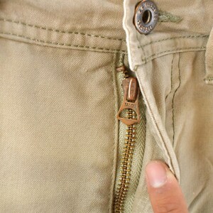 May include: Close-up of khaki-colored pants with a Talon zipper. The zipper is bronze-colored and partially open, revealing the fabric's texture. A button and belt loop are visible.