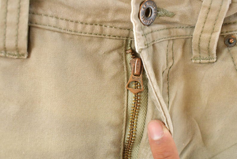 May include: Close-up of khaki-colored pants with a Talon zipper. The zipper is bronze-colored and partially open, revealing the fabric's texture. A button and belt loop are visible.