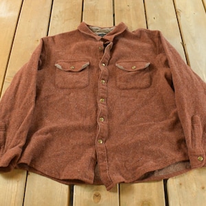 Peut inclure: Une chemise boutonnée brun rougeâtre avec deux poches de poitrine et un col. La chemise à manches longues est faite d'un tissu texturé et possède des boutons dorés.