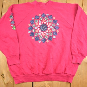Könnte beinhalten: Ein rosafarbenes Sweatshirt mit einem großen, bunten Patchwork-Muster auf der Vorderseite. Das Muster ist ein kreisförmiges Muster aus Quadraten in verschiedenen Blautönen, Rot- und Weißtönen.