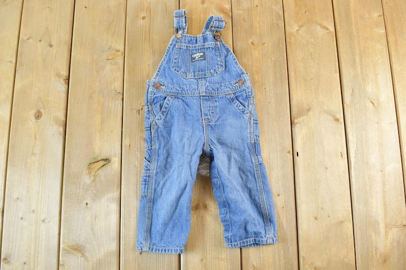 May include: Light blue denim overalls with adjustable straps and multiple pockets. The overalls feature the "OSH KOSH" logo on the bib pocket. A classic, durable garment for children.