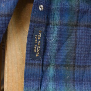 May include: Close-up of a blue, green, and black plaid shirt. The shirt has a snap closure and a tag that reads "HIGH GRADE WESTERN WEAR". The fabric appears to be a soft, brushed material.