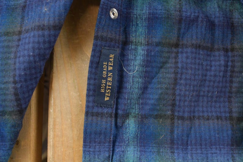 May include: Close-up of a blue, green, and black plaid shirt. The shirt has a snap closure and a tag that reads "HIGH GRADE WESTERN WEAR". The fabric appears to be a soft, brushed material.