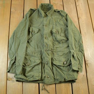 May include: Olive green military-style jacket with multiple front pockets and button closures. The jacket has a collared neckline and long sleeves. A drawstring is visible at the waist. The garment is made of a lightweight fabric.