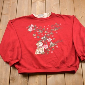 Puede incluir: Sudadera roja con un diseño floral que presenta un gato y pájaros. El diseño está en tonos de rosa, marrón y verde.