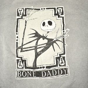 Puede incluir: Sudadera gris con un dise&ntilde;o en blanco y negro de Jack Skellington de The Nightmare Before Christmas. El texto "BONE DADDY" est&aacute; debajo. Las mangas tienen un patr&oacute;n de rayas.