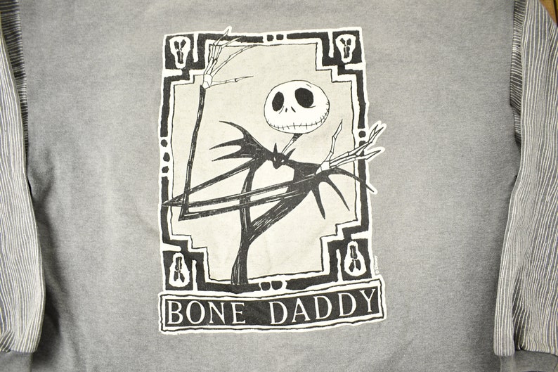 Puede incluir: Sudadera gris con un dise&ntilde;o en blanco y negro de Jack Skellington de The Nightmare Before Christmas. El texto "BONE DADDY" est&aacute; debajo. Las mangas tienen un patr&oacute;n de rayas.