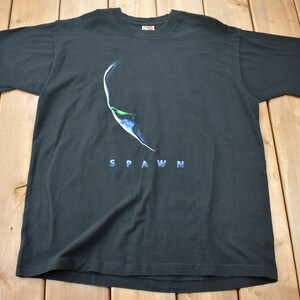 Puede incluir: Camiseta negra con un gráfico azul y verde de la cara de Spawn y la palabra "SPAWN" impresa en azul debajo del gráfico.