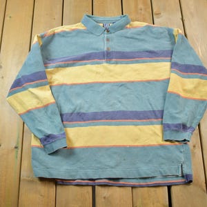 May include: A long-sleeved striped polo shirt with a blue, yellow and purple colour scheme. The shirt has a button-up collar and a single button closure at the neck.