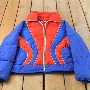 May include: A vintage, quilted jacket in a striking color block design. The jacket features a vibrant red center panel, flanked by blue sleeves and side panels. White piping outlines the red sections. The jacket has a high collar and a full-length zipper.