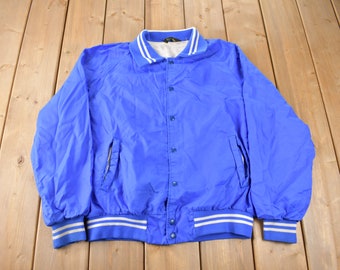 May include: A blue nylon varsity jacket with white and blue striped trim around the collar, cuffs, and waistband. The jacket has a button closure and two front pockets.